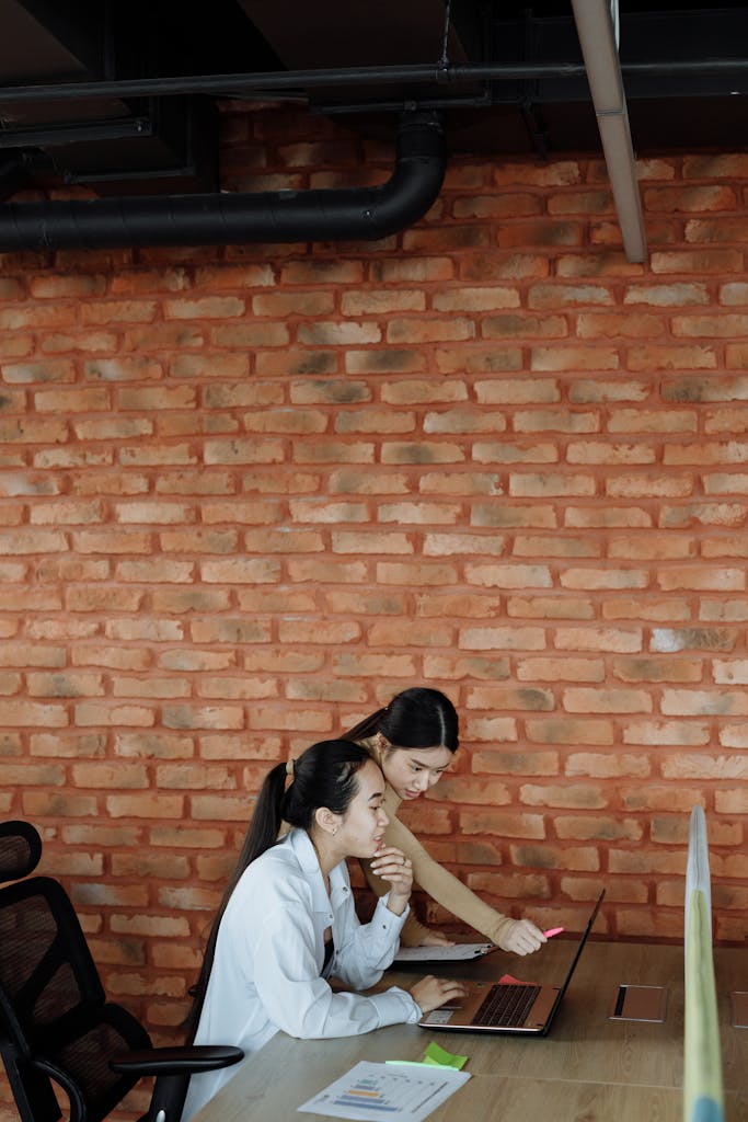 Asian women collaborating on a project using a laptop in a modern brick-walled office setting.